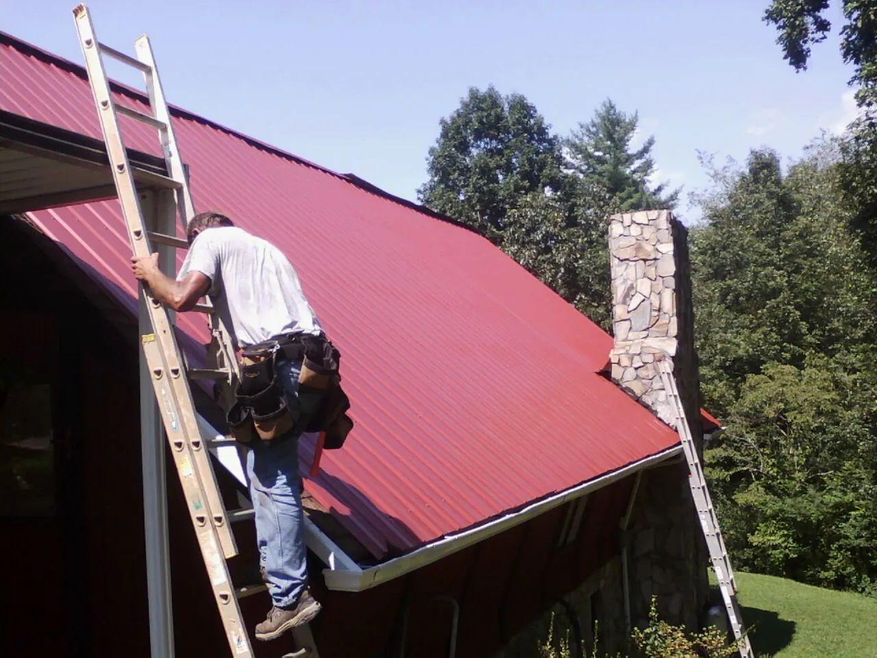 Roofer on a ladder beside a red metal roof and stone chimney — Soffit & Fascia Repair by Roofing Brothers in Tariffville